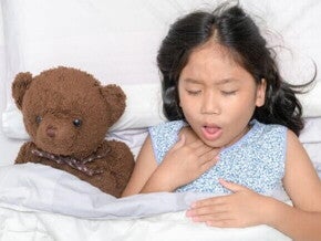 Uma menina deitada na cama com expressão de desconforto, segurando a garganta, ao lado de um ursinho de pelúcia.
