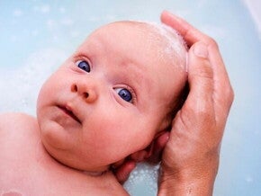 Um close-up de um bebê recém-nascido, deitado na água do banho com espuma. Uma mão adulta com sabão na ponta dos dedos está suavemente lavando a cabeça do bebê.