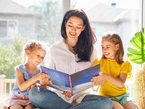 Uma mulher sorridente está sentada lendo um livro para duas meninas pequenas sentadas ao seu lado.