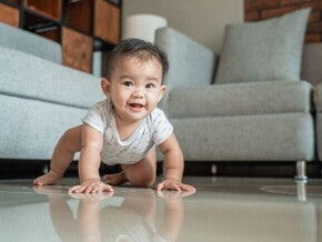 Um bebê sorridente engatinhando em um piso brilhante, com um sofá cinza e uma parede de tijolos ao fundo, ilustrando um marco de desenvolvimento motor. Um bebê sorridente engatinhando em um piso brilhante, com um sofá cinza e uma parede de tijolos ao fundo, ilustrando um marco de desenvolvimento motor.