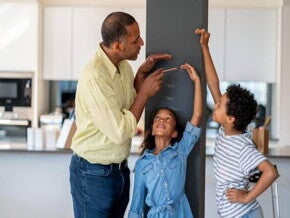 Em uma cozinha, está um homem, uma menina e um menino. A menina está encostada em uma parede, enquanto o homem e o menino marcam na parede qual é a altura dela. Em uma cozinha, está um homem, uma menina e um menino. A menina está encostada em uma parede, enquanto o homem e o menino marcam na parede qual é a altura dela.