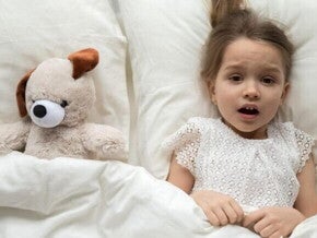 Uma menina assustada está deitada na cama, coberta por um lençol, com expressão de medo, ao lado de um cachorro de pelúcia.