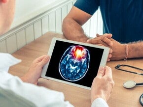Um médico de jaleco branco mostra um tablet com uma imagem de ressonância magnética de um cérebro para um paciente. A imagem ilustra as graves consequências de doenças como a meningite B.