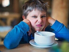 Meu filho não quer comer Um menino fazendo uma expressão de desgosto, está apoiando o rosto nas mãos e olhando para a câmera. Na frente dele, em uma mesa de madeira, há uma tigela branca em um pires.