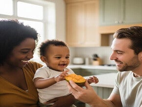 Mãe e pai interagem com bebê sorridente, oferecendo frutas, em ambiente iluminado.