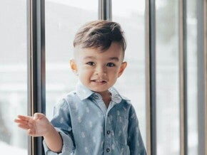 Menino sorrindo com uma camisa azul, com uma porta de vidro ao fundo.