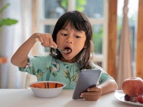 Uma menina come enquanto olha para um celular na mesa.