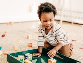 Criança pequena brincando no chão com blocos de montar coloridos em uma bandeja verde, com vários brinquedos espalhados ao redor sobre um tapete bege em um ambiente iluminado.
