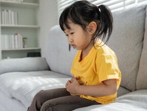 Uma menina está sentada em um sofá, segurando a barriga com as duas mãos, com uma expressão de desconforto ou dor.
