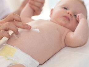 Uma mão de adulto gentilmente aplica creme na barriga de um bebê deitado, destacando o cuidado com a pele delicada do bebê.