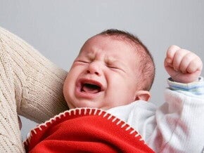 Um bebê chorando intensamente, enrolado em um cobertor vermelho, com o rosto virado para cima, demonstrando grande desconforto.