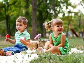 Um menino e uma menina estão sentados em uma toalha em um parque. Sobre ela temos algumas cestas e lanchinhos. O cenário todo remete muito a um dia de piquenique