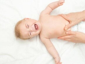 Bebê deitado de costas em uma superfície branca, chorando, enquanto um adulto realiza uma massagem suave em sua barriga.