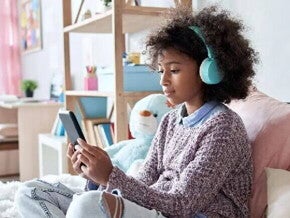 #ParaTodosVerem Menina negra pré-adolescente, com cabelo cacheado, solto, usando fones de ouvido azuis enquanto olha a tela do celular. Ela está sentada em uma cama com almofadas e veste blusão com calça jeans. O fundo da foto é um quarto de menina.