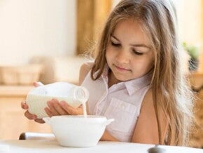 Menina sentada na mesa de jantar despejando leite em um tigela de cereais Menina sentada na mesa de jantar despejando leite em um tigela de cereais