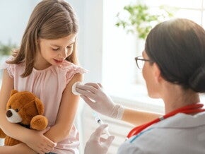 Uma menina segura um ursinho de pelúcia enquanto uma profissional de saúde, prepara o braço da menina para uma injeção com um algodão.