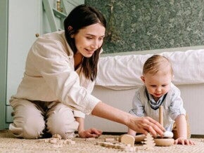 Mulher sorridente ajoelhada no chão de um quarto brincando com seu bebê.