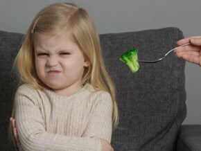 Uma menina pequena está sentada em um sofá cinza com os braços cruzados e uma expressão de desagrado ou nojo no rosto, franzindo as sobrancelhas e a boca. 