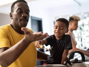 Pai e filho cozinham juntos como uma homenagem de dia dos pais. O pai está mexendo uma panela e o filho, ao seu lado, o ajuda. 