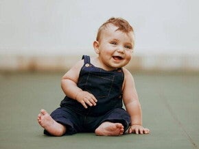 Bebê pequeno, de bochechas rechonchudas e olhos azuis, sentado em um chão plano e sorrindo, vestindo um macacão jeans azul-marinho.