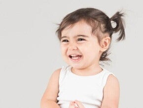 Close-up de um bebê sorrindo, com o cabelo preso em um rabo de cavalo no topo, vestindo uma regata branca, com fundo cinza-claro.