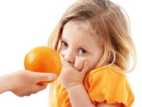 Uma mão oferece uma laranja a uma menina, que cobre a boca com a mão e olha para a câmera com uma expressão de repulsa ou aversão.