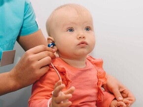Profissional da saúde aplicando o teste da orelhinha em um bebê no colo, usando um pequeno fone no ouvido da criança para verificar a capacidade auditiva.