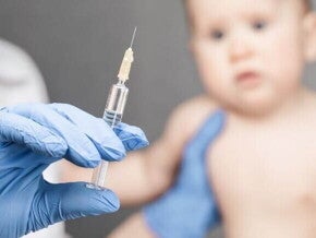 Mão com luva cirúrgica segurando seringa com a vacina da coqueluche, enquanto bebê aguarda a aplicação