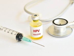 Vacins HPV nonavalente Vista superior de um frasco de vacina rotulado "HPV vaccine" com líquido amarelado, colocado entre uma seringa hipodérmica e um estetoscópio médico, todos dispostos sobre uma superfície branca.