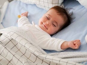 Bebê dormindo de barriga para cima em uma cama com lençóis e cobertor, transmitindo sensação de conforto e tranquilidade.