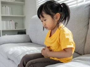 Uma criança sentada no sofá com a mão na barriga, expressando dor. A imagem representa o mal-estar causado por verme infantil.
