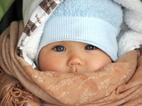 Um close-up de um bebê usando um chapéu azul e um cobertor marrom enrolado em volta do rosto, mostrando como agasalhar um bebê para o frio e como vestir um bebê de acordo com a temperatura. Um close-up de um bebê usando um chapéu azul e um cobertor marrom enrolado em volta do rosto, mostrando como agasalhar um bebê para o frio e como vestir um bebê de acordo com a temperatura.