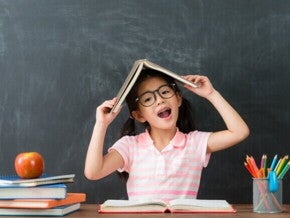 Uma menina com óculos, sentada à mesa, faz uma pose divertida com um livro aberto sobre a cabeça. Ela sorri com a boca aberta em frente a um quadro negro, com um monte de livros e lápis de cor ao seu lado. A imagem é lúdica e representa a criatividade e a diversão do aprendizado, remetendo ao clima de volta às aulas.