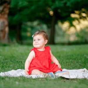 Sua criança completou um ano? Preparar, explorar, descobrir! Criança de um ano com vestido vermelho e fita no cabelo posa para foto em um parque, sentado em um pequeno cobertor colocado sobre a grama.