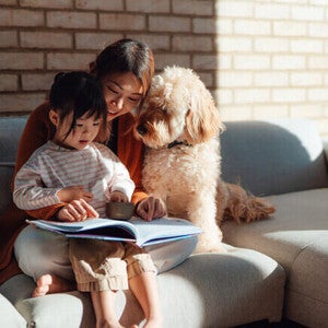 Pessoa lendo um livro ilustrado para uma criança enquanto estão sentadas no sofá com o cachorro da família