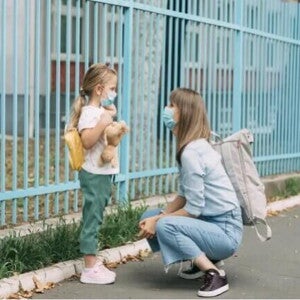 Uma mãe está agachada em frente a sua filha, perto de um portão. A menina está de mochila, olhando para a mãe e segurando um urso de pelúcia. Uma mãe está agachada em frente a sua filha, perto de um portão. A menina está de mochila, olhando para a mãe e segurando um urso de pelúcia.