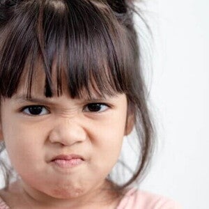 Close do rosto de uma menina expressando agressividade na primeira infância. Ela está com as sobrancelhas franzidas, nariz contraído e lábios pressionados em um sinal nítido de raiva ou frustração. O fundo é neutro e claro, destacando a expressão emocional da criança.