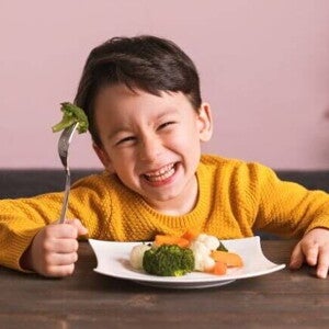Menino sorridente segurando um garfo com brócolis e um prato cheio de vegetais cozidos à frente, representando alimentos para aumentar a imunidade de forma divertida e nutritiva.