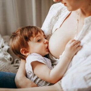 Close-up de uma mãe em uma camisa branca amamentando seu filho (cerca de 1 a 2 anos) dentro de casa.