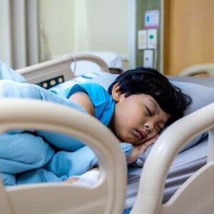 Uma criança dormindo profundamente em uma cama de hospital, enrolada em lençóis azuis, em um ambiente hospitalar.