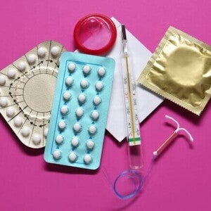 Visão aérea de vários métodos contraceptivos, incluindo cartelas de pílulas anticoncepcionais, preservativos (camisinhas), um DIU (dispositivo intrauterino) e um termômetro, dispostos sobre um fundo rosa.