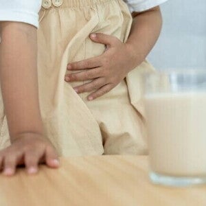 Uma criança está segurando a área da barriga com uma das mãos, indicando dor ou desconforto abdominal. Um copo com um líquido que se assemelha ao leite está desfocado em primeiro plano sobre uma mesa.