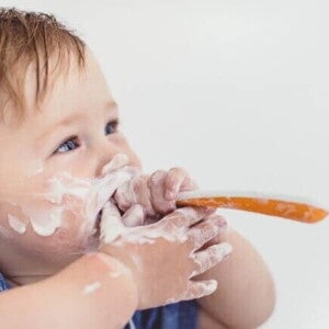 Close-up de um bebê, com o rosto e as mãos sujos de iogurte ou creme branco, segurando uma colher laranja e branca e olhando para cima.