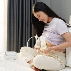 Uma mãe sorridente e sentada na cama utilizando uma bomba elétrica para extrair e coletar o leite materno em frascos.