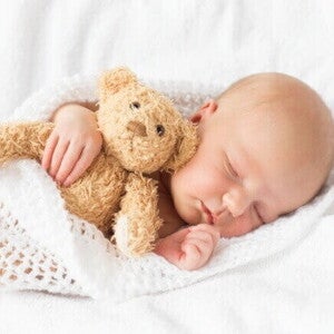 Bebê dormindo tranquilamente enrolado em uma manta branca e abraçado em um urso pequeno de pelúcia.