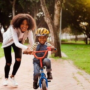 Bicicleta infantil: qual a melhor idade para começar e quais os modelos ideais? Foto de um menino sorrindo, em uma bicicleta sem rodinhas e ao lado uma mulher adulta também sorrindo segurando em um dos lados do guidão da bicicleta.