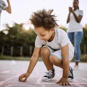 Imagem de uma criança ao centro brincando de uma brincadeira africana no chão. Ao fundo seus pais torcem por ela Imagem de uma criança ao centro brincando de uma brincadeira africana no chão. Ao fundo seus pais torcem por ela