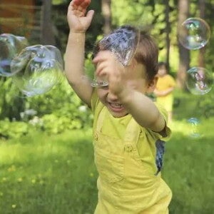 Brincadeiras ao ar livre Um menino sorri com as mãos para o alto enquanto passa por bolhas de sabão em um ambiente externo arborizado, com outro menino ao fundo. A foto faz alusão ao tema de brincadeiras ao ar livre.