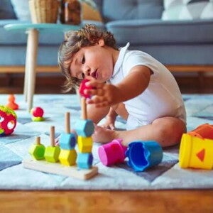 Um bebê está sentado no tapete de uma sala de estar mexendo com brinquedos de empilhar e montar, que é uma das brincadeiras que estimulam o desenvolvimento infantil