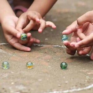 Duas crianças brincam de bola de gude no chão em um círculo desenhado de giz. A foto faz alusão ao tema de brincadeiras indígenas Duas crianças brincam de bola de gude no chão em um círculo desenhado de giz. A foto faz alusão ao tema de brincadeiras indígenas